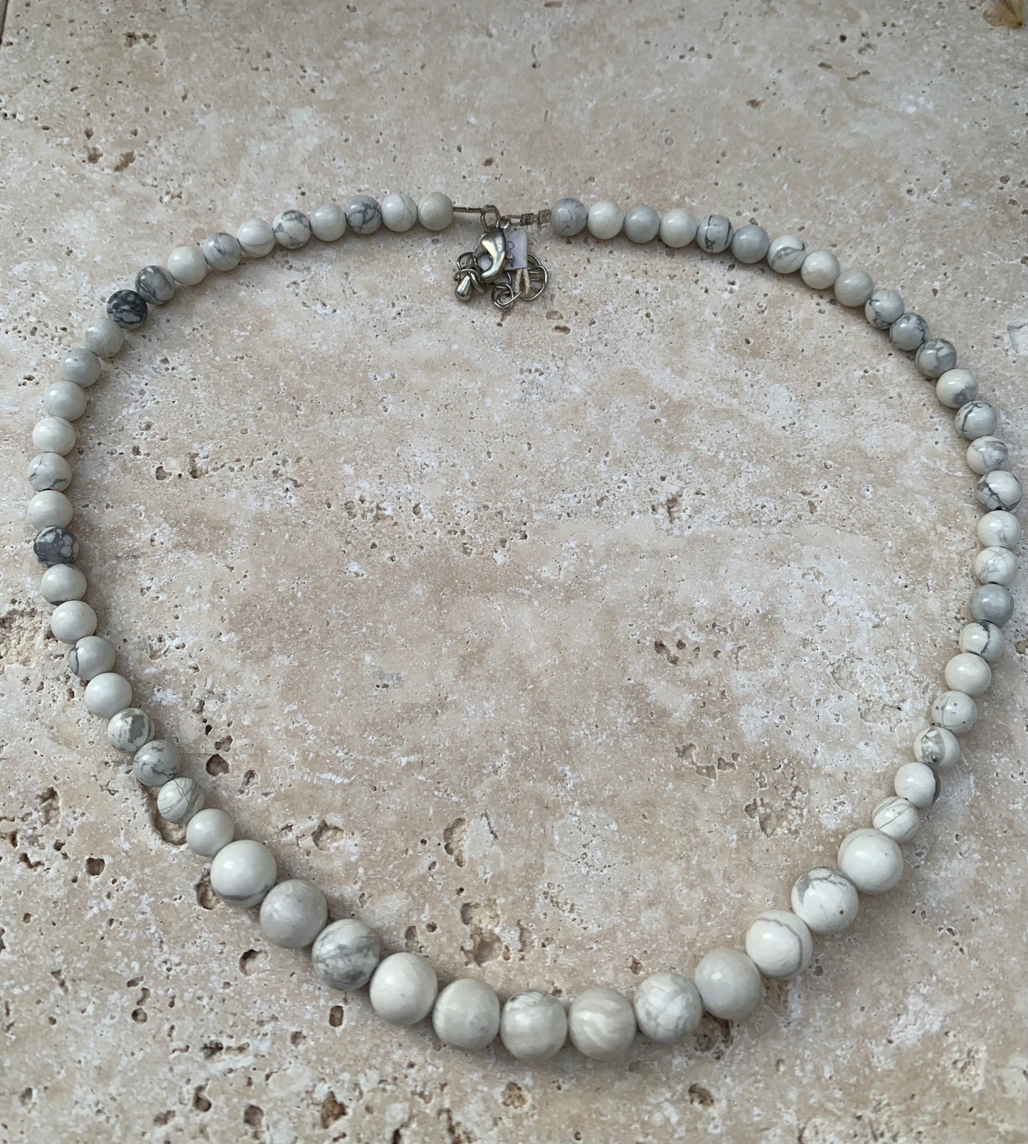 Howlite, 'collier/ chute de perle', de forme ronde de 4 à 10 millimètres de diamètre. 44 centimètres de longueur + 4 cm de rajout en chaînette. Fermoir de type 'mousqueton'. Très belle pièce !