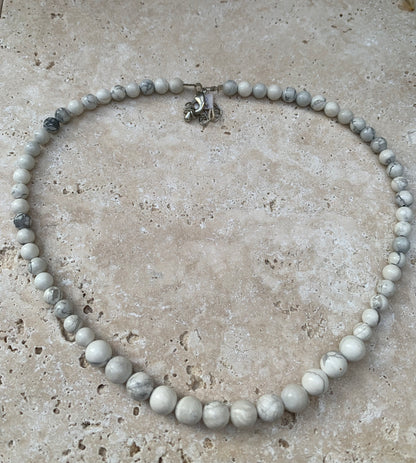 Howlite, 'collier/ chute de perle', de forme ronde de 4 à 10 millimètres de diamètre. 44 centimètres de longueur + 4 cm de rajout en chaînette. Fermoir de type 'mousqueton'. Très belle pièce !